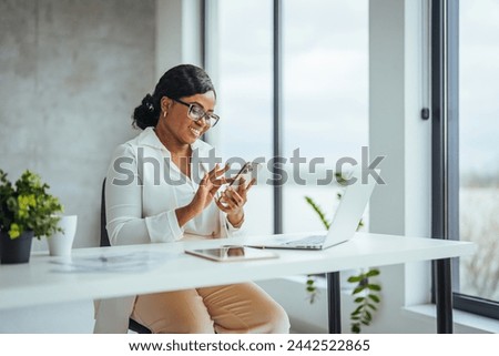 African black business woman using smartphone while working on laptop at office. Smiling african american businesswoman lookingup at copy space while working on phone.