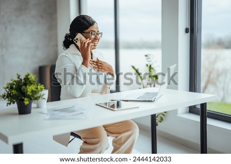 African black business woman using smartphone while working on laptop at office. Smiling african american businesswoman lookingup at copy space while working on phone. Successful woman entrepreneur