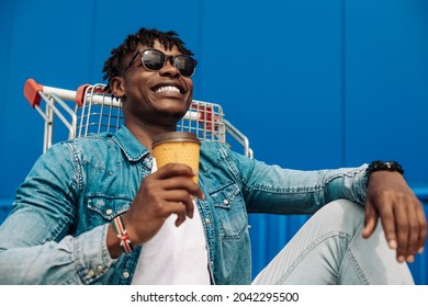 African black man holding a cup of coffee or tea in a paper cup in his hand, sitting on a shopping trolley, near a shopping center, against the background of a blue building - Powered by Shutterstock