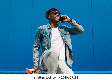 African Black Man Sitting On A Shopping Cart Talking On Phone, Making A Phone Call, Shopping Online Talking To An Operator, Against The Background Of A Blue Building