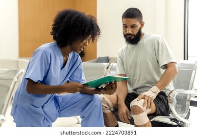 African black nurse woman asking injured informations from young Caucasian patient man in hospital, female afro hair nurse in blue hospital uniform checking beard man patient about injured wound