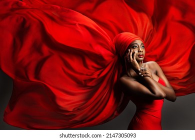 African Black Woman In Red Silk Headscarf. Fashion Dark Skinned Model Portrait In Turban With Golden Jewelry In Hijab Flying Chiffon Fabric Over Gray Background. Stylish Arab Beauty Women Headwear