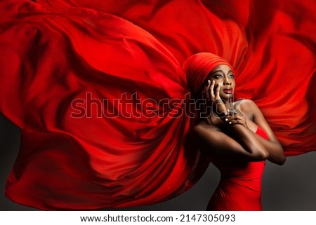 African Black Woman in Red Silk Headscarf. Fashion Dark Skinned Model Portrait in Turban with Golden Jewelry in Hijab flying Chiffon Fabric over Gray Background. Stylish Arab Beauty Women Headwear