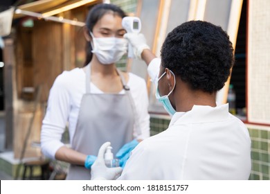 African Black Woman Retail Business Staff Or Grocery Shop Keeper Checking Fever Of Asian Coworker By Portable Thermo Scan Device; Concept Of New Normal Business Practice