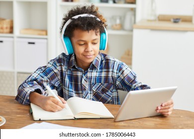 African boy in headphones using digital tablet and making notes in his notebook while sitting at the table at home - Powered by Shutterstock