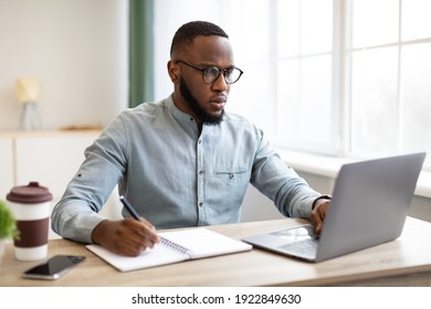 African Business Guy Working On Laptop Taking Notes Sitting At Workplace Indoors. Male Office Worker Writing At Work. Business Career And Corporate Lifestyle Concept. Selective Focus