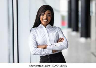 African Business Woman Looking Into The Camera With Her Arms Crossed While Standing In Front Of Large Glass Windows With A Cityscape Behind Her.