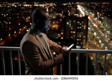 African businessman in formalwear looking through contacts in mobile phone while standing on balcony in the night