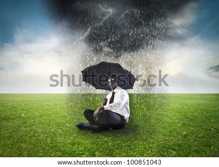 African businessman sitting under an umbrella on a green meadow with stormy cloud over him