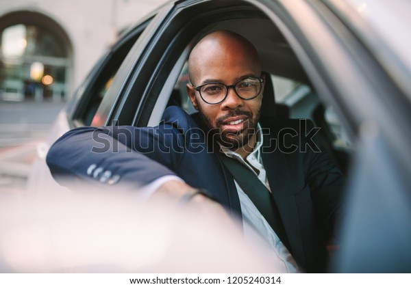 African businessman in suit peeking out of car\
window and looking at camera while driving. Handsome businessman in\
suit driving car on city\
road.