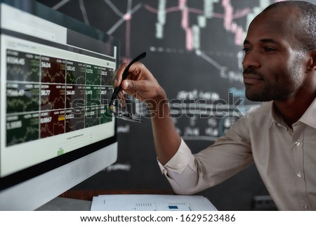 African businessman, trader sitting in front of computer screen and holding glasses while looking at graph chart. Blackboard full of data analyses in the background. Stock trading, people concept.