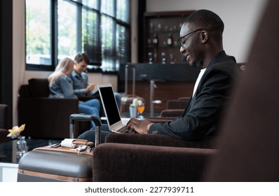 African businessman using laptop while sitting in airport lounge with blurred couple in background. Business travelers waiting for their flight at airport terminal. - Powered by Shutterstock