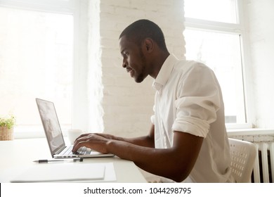African businessman working on laptop sitting at home office desk, happy black intern employee looking at computer screen typing or browsing web, using software application for business and education