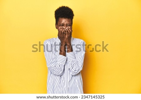 African businesswoman, blue striped shirt, yellow backdrop, laughing about something, covering mouth with hands.