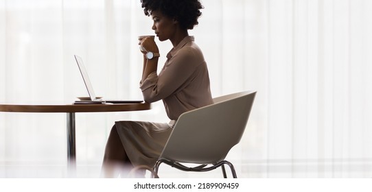 African Businesswoman Drinking Coffee And Looking At Laptop On Table. Side View Of Female Executive Using Laptop And Having Coffee At Office.