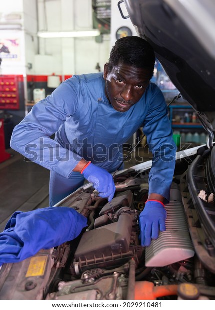 African car mechanic with screwdriver\
repairing car engine under the hood in auto\
service