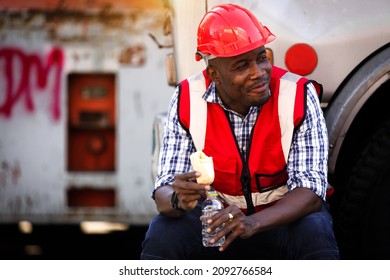An African cargo truck driver or maintenance  engineer in red protective clothing and a hard hat sits down to rest, eats snacks and water during breaks with a smiling face.
