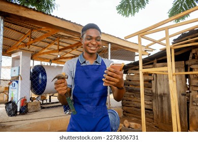 african carpenter holding a phone and hammer feeling very excited wearing an apron working - Powered by Shutterstock