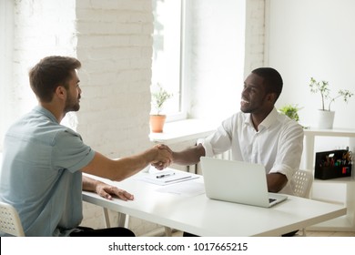 African and caucasian businessmen shaking hands over office desk, satisfied white partner closing successful business deal with black investor, handshake after making agreement or getting hired