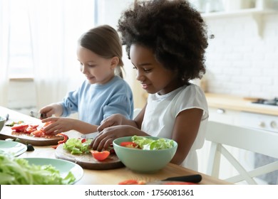 African and Caucasian little girls best friends cooking together in modern kitchen. Multiracial cousins hold knives cutting vegetables on wooden board prepare healthy salad making surprise for parents - Powered by Shutterstock