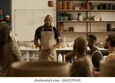 African ceramist wearing an apron talking with a diverse group of coworkers during a meeting together in a ceramics studio - Powered by Shutterstock