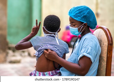 African Child With Face Mask Tied Behind Her And Sitting On A Health Care Provider Or Doctor With Hands Raised Up Surety,physician Auscultating Back Of Child And Wearing Face Mask For Protection