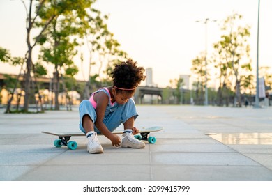 African Child Girl Kid Tying Sneaker Shoes While Skateboarding At The Park. Cute Preschool Girl Enjoy And Having Fun Outdoor Lifestyle Practicing Extreme Sport Longboard Skating On Summer Vacation.