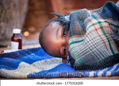 African Child Toddler Sick With Malaria Medicine In The Background, Laying Down On A Blanket In The Yard