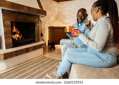 African couple drinking hot chocolate in front of cozy fireplace at home - Focus on man face - Powered by Shutterstock