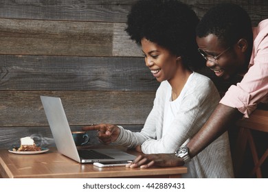 African Couple Having Coffee At A Cafe: Black Girl Sitting In Front Of Laptop Pointing At The Screen And Laughing, Looking Through Wedding Photos Together With Her Husband Who Is Standing Next To Her