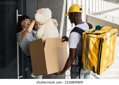 African Courier Bring Parcel To Little Girl With Mom At The Door Of Their House. Opening Parcel, Happy Child Takes Out Of Box Fluffy Teddy Bear Toy, Rejoices In The Surprise, Family Delivery.