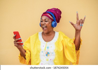 African Crazy Senior Woman Wearing Traditional African Dress While Listening Music On Mobile Phone App - Party Concept - Focus On Face
