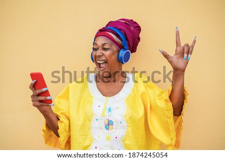 African crazy senior woman wearing traditional african dress while listening music on mobile phone app - Party concept - Focus on face