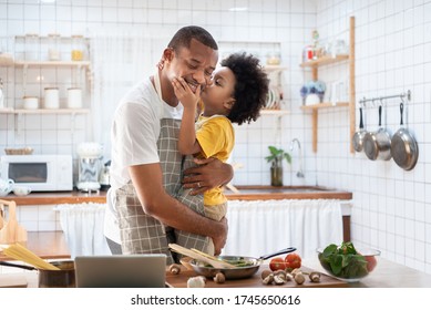 African Cute Little Boy In Yellow Casual Kissing Dad While Cooking At Home Together. Happy Smiling African-American Father While Hug And Carry His Son In Kitchen. Joyful Black Family, Love Emotion.