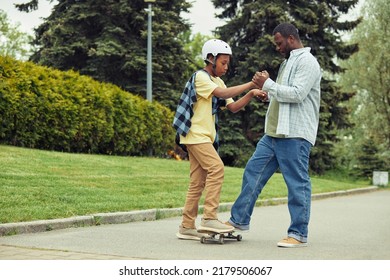 African Dad Helping His Son To Stand On Skateboard And Teaching Him To Ride Outdoors In Park