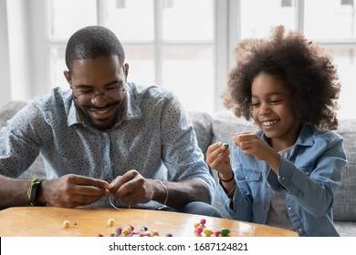 African dad and little daughter sit on couch use colorful beads making bracelet jewellery enjoy pastime together at home activity promote development, fine hand motor skills, pastime and hobby concept