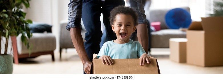 African dad play with excited toddler son riding seated inside of box in moving day at new home, little kid enjoy game with daddy, relocation concept. Horizontal photo banner for website header design