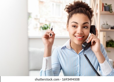 African Descent Female Tour Agent Sitting At Table In Office Answering Landline Phone Call Communication With Client Holding Pen Smiling Thoughtful Close-up