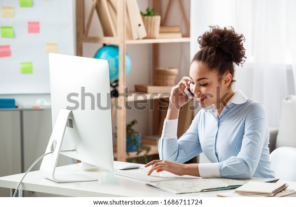 African descent female tour agent sitting\
at table in office answering phone call talking with client looking\
information on computer smiling\
joyful