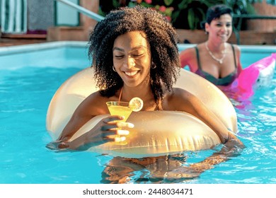 African descent woman with a bright smile relaxes on a golden inflatable ring in a pool, sipping a refreshing lemon cocktail on a sunlit day, with a friend in the background. - Powered by Shutterstock