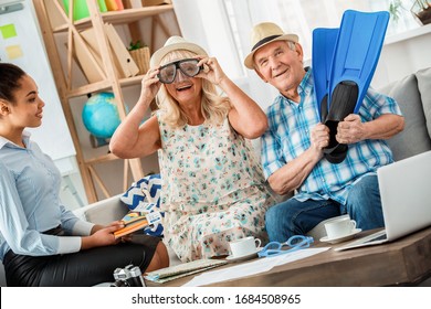 African Descent Woman Travel Agent At Office With Senior Couple Wearing Beach Hats Holding Swimming Mask And Flippers Looking Camera Smiling Joyful