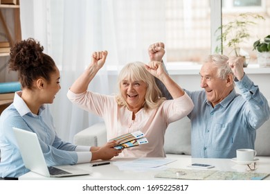 African Descent Woman Travel Agent At Office Giving Passports And Plane Tickets To Senior Couple Holding Hands Up Screaming Smiling Cheerful