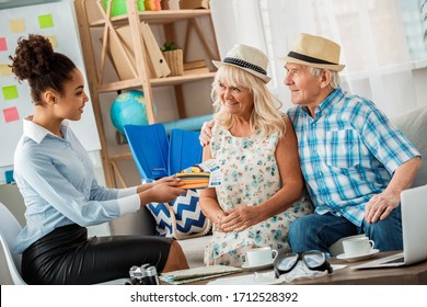 African descent woman travel agent at office smiling happy giving passports and plane tickets to senior couple sitting on sofa wearing beach hats side view - Powered by Shutterstock