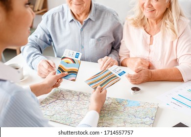 African Descent Woman Travel Agent At Office Sitting At Table Giving Passports In Colorful Cover And Plane Tickets To Senior Couple Smiling Happy Close-up