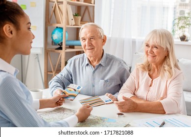 African Descent Woman Travel Agent At Office Sitting At Table Giving Plane Tickets And Passports To Senior Couple Smiling Joyful Side View