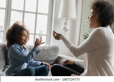 African disabled daughter and her mother sitting on sofa show symbols with hands using visual-manual finger gestures interacting at home. Hearing loss deaf person sign language learning school concept