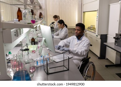African disabled scientist sitting in wheelchair at the table and working on computer with his colleague in the background in the lab