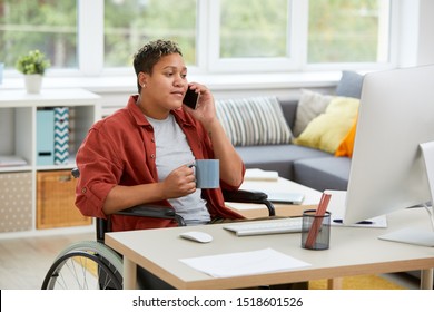 African disabled woman sitting in wheelchair at the table and looking at computer monitor while talking on mobile phone and drinking coffee at home - Powered by Shutterstock