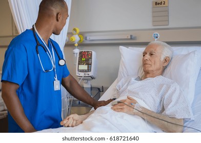 African Doctor Checking Blood Pressure On Monitor Screen For Patient Health Status. Male Nurse Doing Routine Checkup Of Senior Patient In Hospital Room.