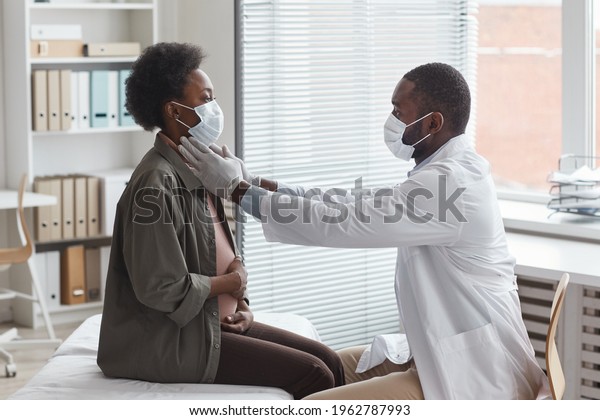 African doctor in protective mask\
examining pregnant woman during her visit at doctor\'s\
office